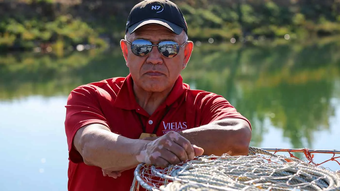 Man in sunglasses and a hat leaning on fishing gear looking stern at camera.