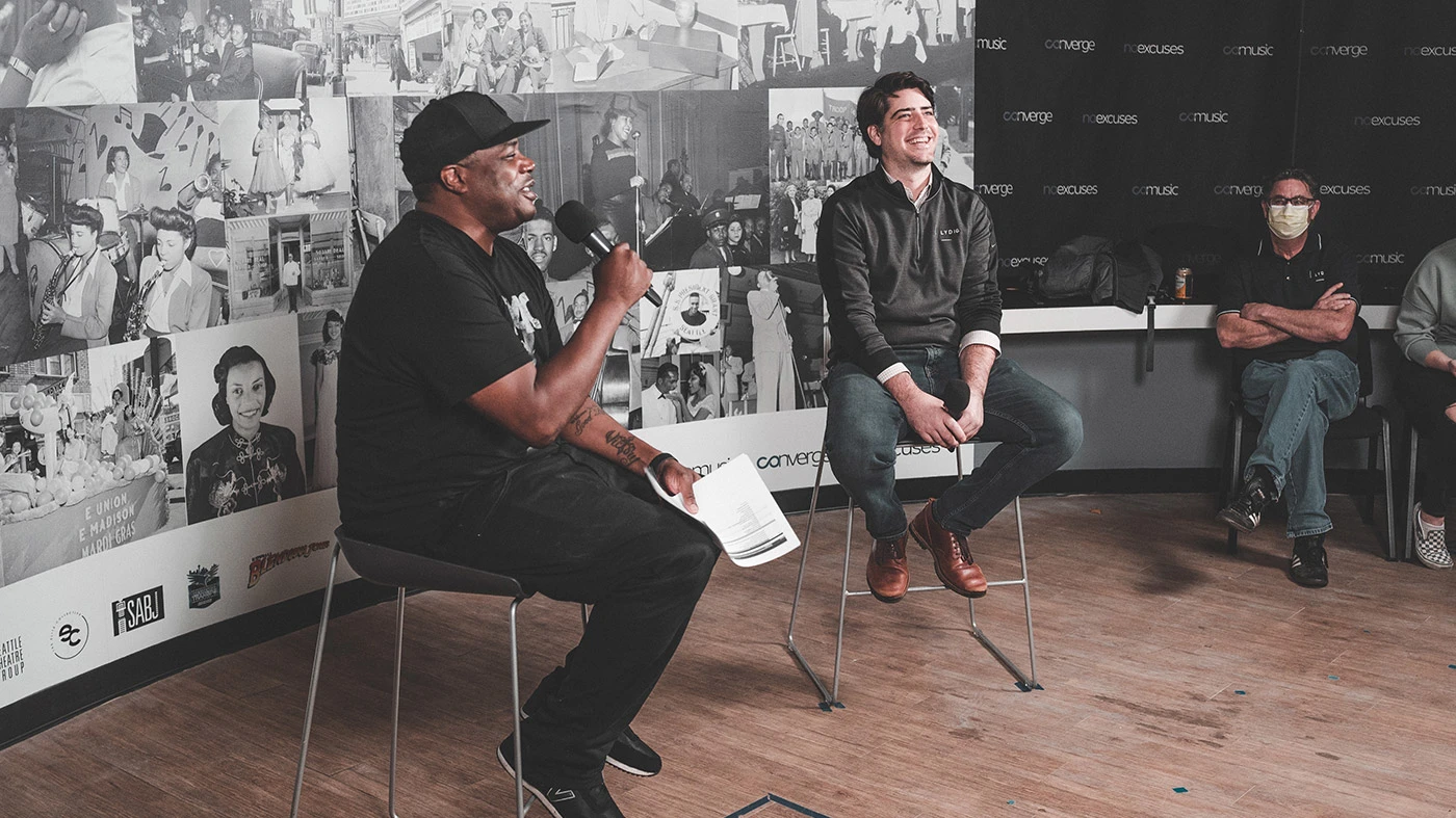 Two men in black sitting on stools talking, in front of black in white picture collage.
