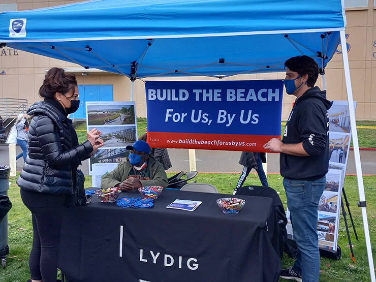 Two people standing next to a Lydig information table underneath a blue canopy with a sign with the text Build the Beach, For Us by Us.