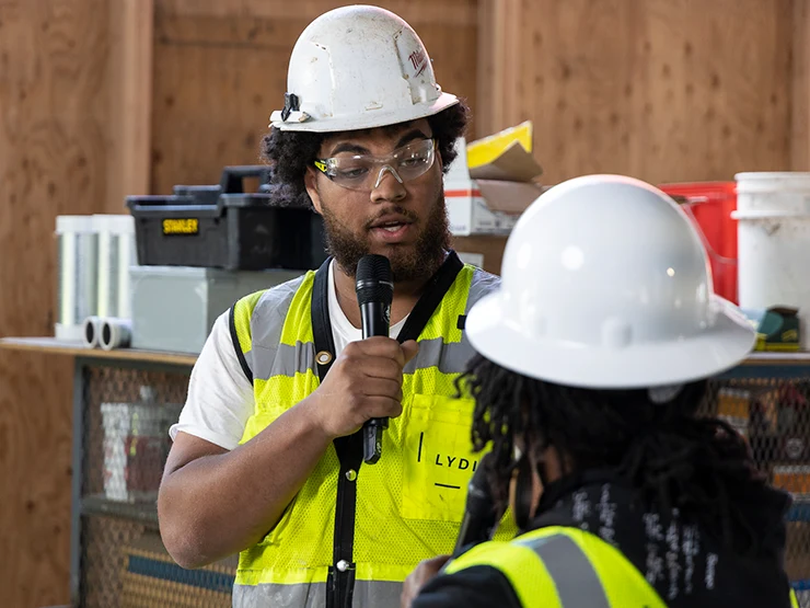Close up of man in construction hard hat and vest talking to another person in a construction hard had and vest.