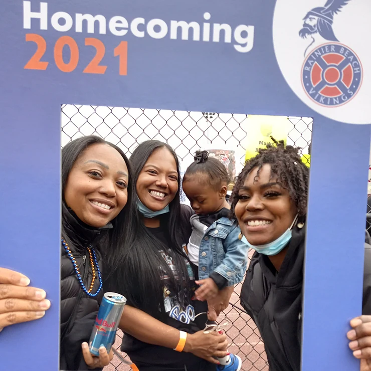 Three women, one holding a baby, taking a picture holding a blue cut out frame with text “Homecoming 2021 hashtag Build The Beach For Us By Us