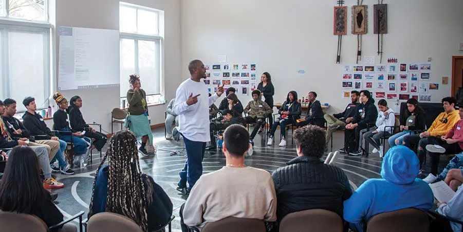 Large group of people in a room sitting around in a circle, listening to man, who is standing, speak.