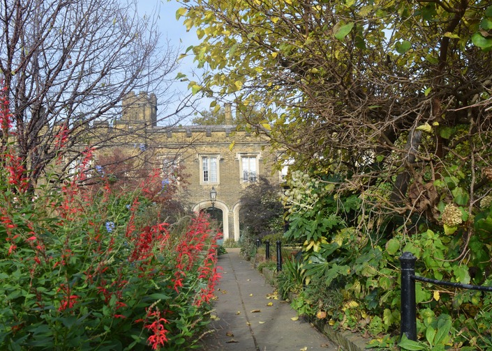A Victorian building with a design featuring an arcade and decorative battlements and turret visible between trees which have largely lost their leaves, foliage and red flowers.