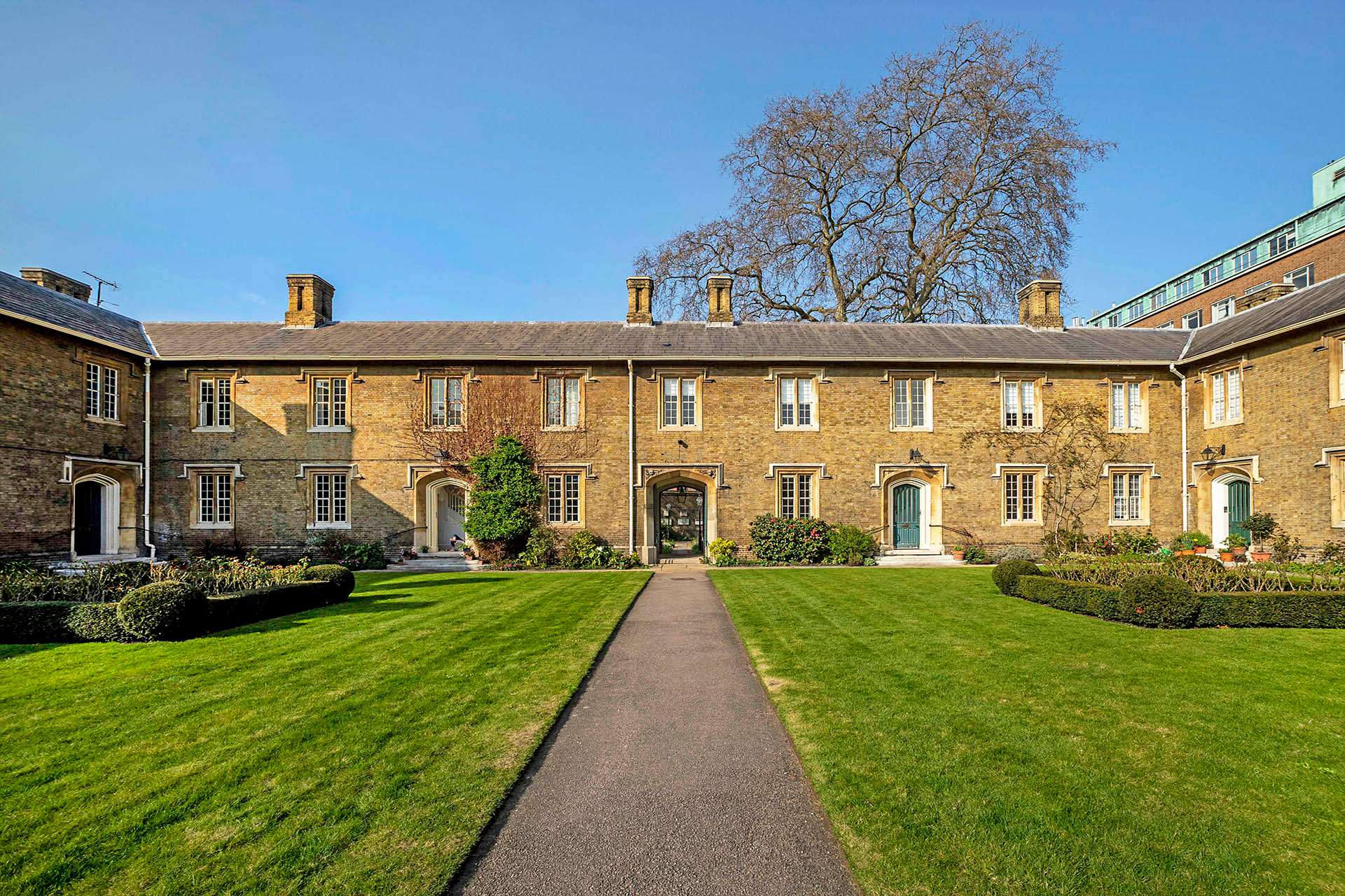 Yellow-brick Victorian courtyard housing with green lawns and topiary flowerbeds in the foreground and a blue sky and leafless tree behind