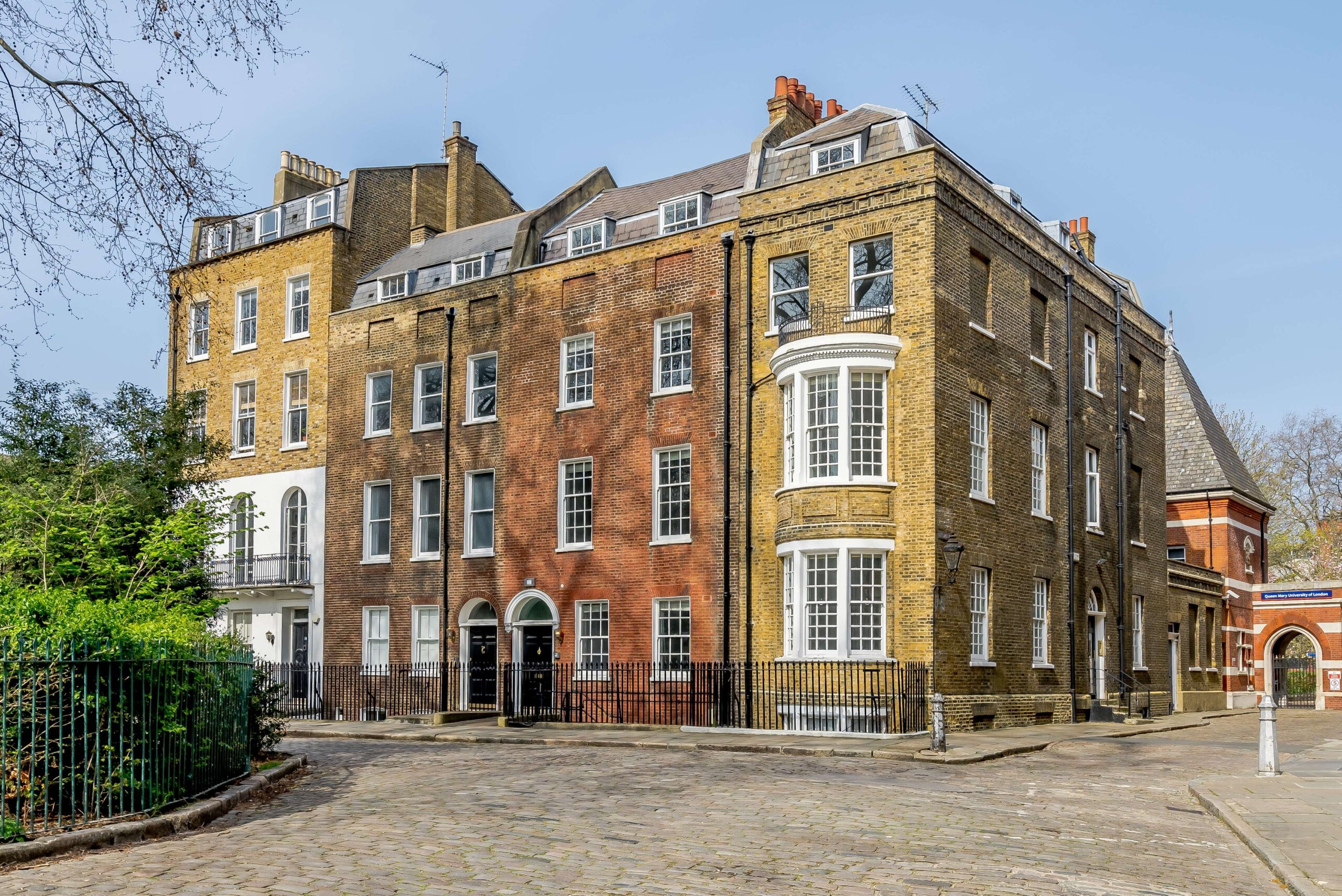 Four large multi-story red and yellow Victorian terrace houses, one with round bay windows, with a cobbled road in the foreground and greenery of a garden square with iron railings just visible.