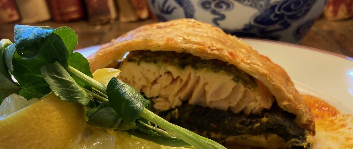 a white plate with a slice of salmon en croute, lemon wedges and watercress with the base of some books and a blue and white lamp just visible behind