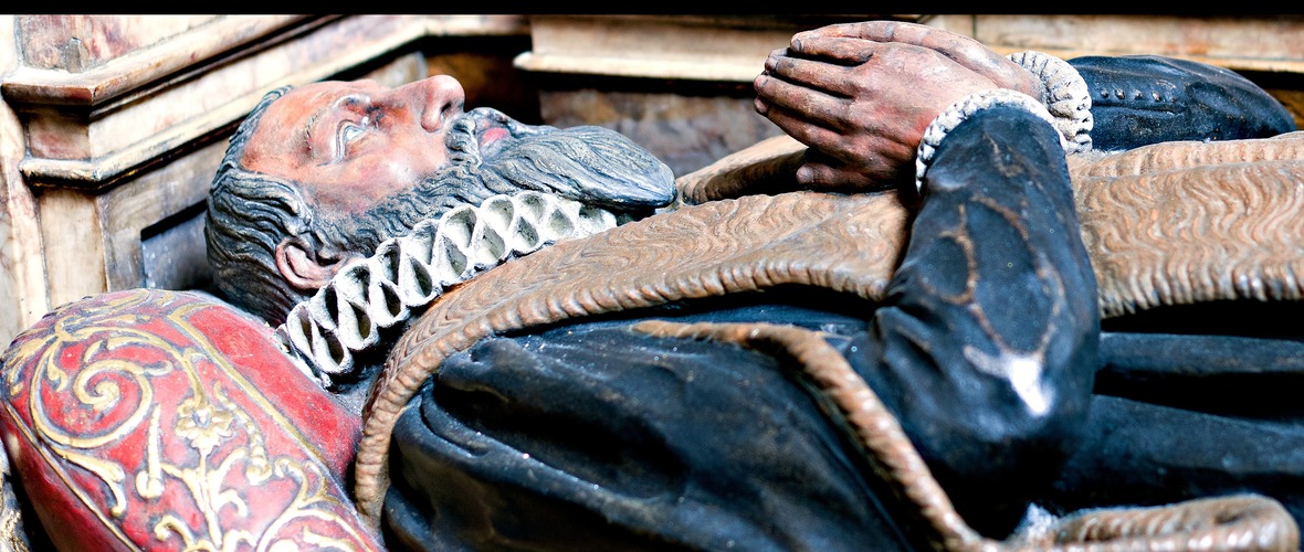 effigy of a Tudor man with a beard, white ruff and black fur-trimmed cloak lying on a red patterned pillow wiht his hands held in prayer
