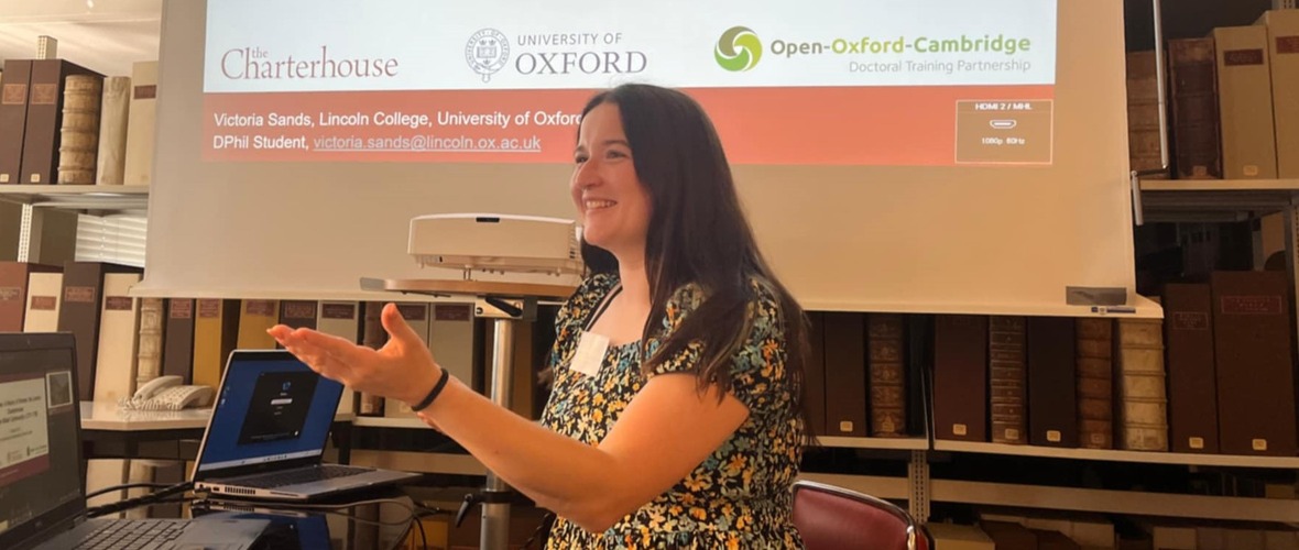 Victoria Sands - a young woman with long brown hair and a floral dress - seated infront of shelves of books, a projector and drop down screen with the Charterhouse, University of Oxford and Open-Oxford-Cambridge visible