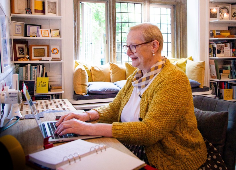 Woman wearing a mustard yellow cable knit cardigan in side profile, using a laptop at a table with a large open leaded window behind her with a window seat containing yellow cushions