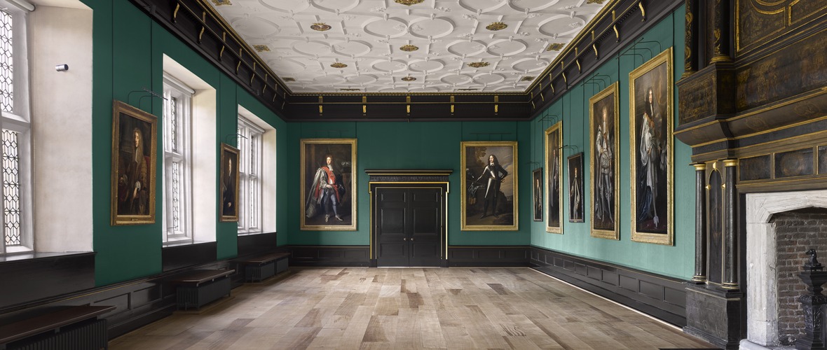 View towards the doorway in a historic room with a white and gold moulded ceilng, green silk walls and seventeenth century portraits hung on the walls