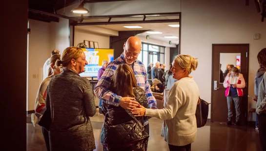 A group of people chat warmly in a building lobby. In the center, a smiling man in a plaid shirt gives a welcoming hug to a woman.