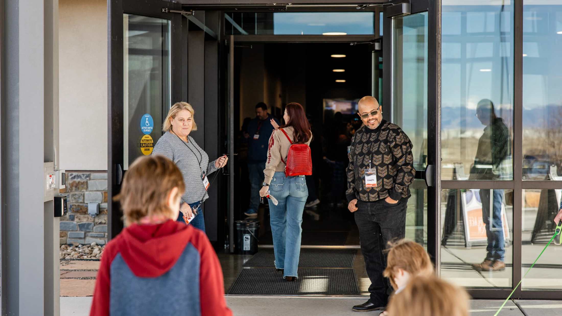 A man and woman stand at the glass doors of a building, greeting people as they arrive. A woman with a red backpack walks inside.