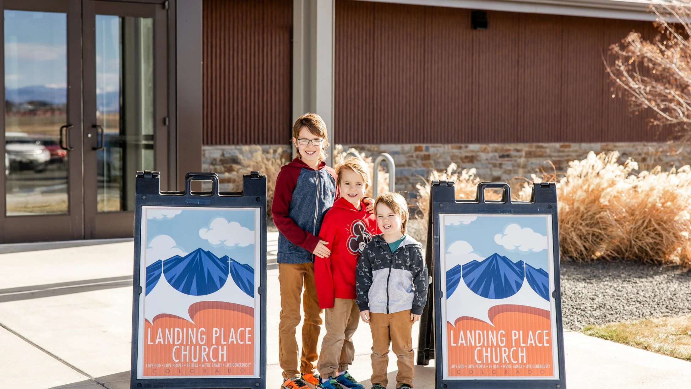Three young boys smile for a photo while standing between two A-frame signs for Landing Place Church in front of the building's entrance.