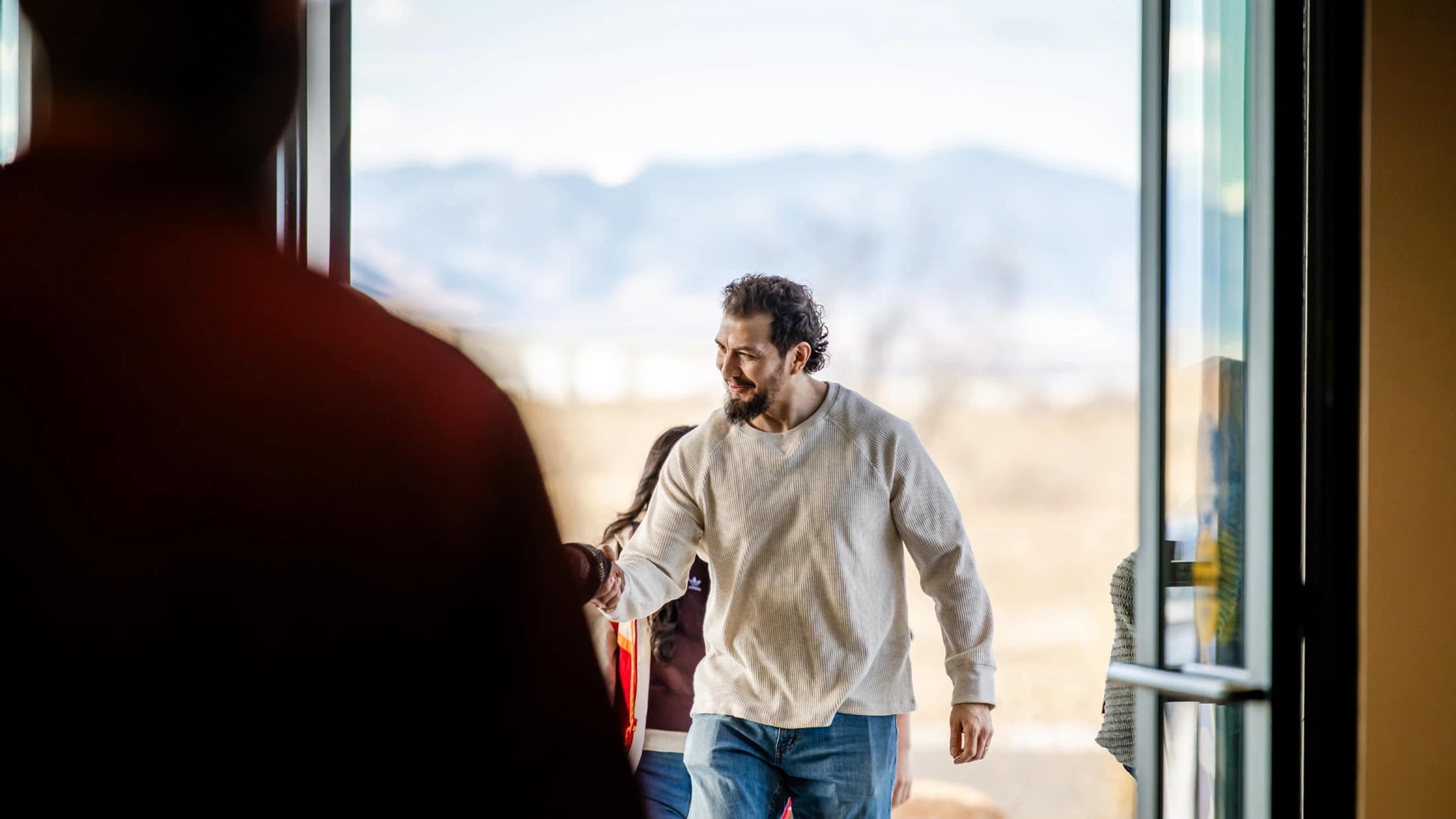 A smiling man with a beard shakes hands with someone in a doorway, with a view of mountains in the background. The person he is greeting is in the foreground.