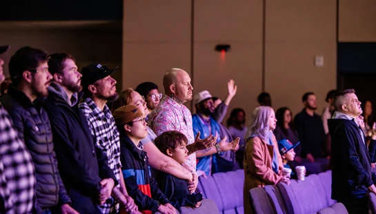 A diverse group of people stands in an auditorium with purple lighting. Many look on with reverence, and some raise their hands in worship.