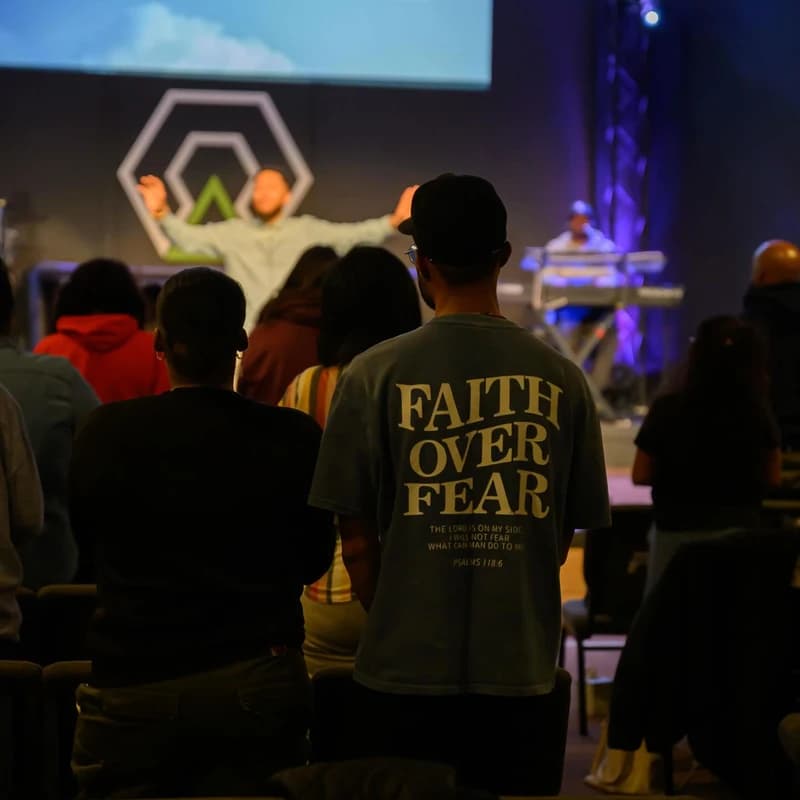 View from behind of a congregation during a church service. A man in the foreground wears a t-shirt that reads 'Faith Over Fear' with a Bible verse.