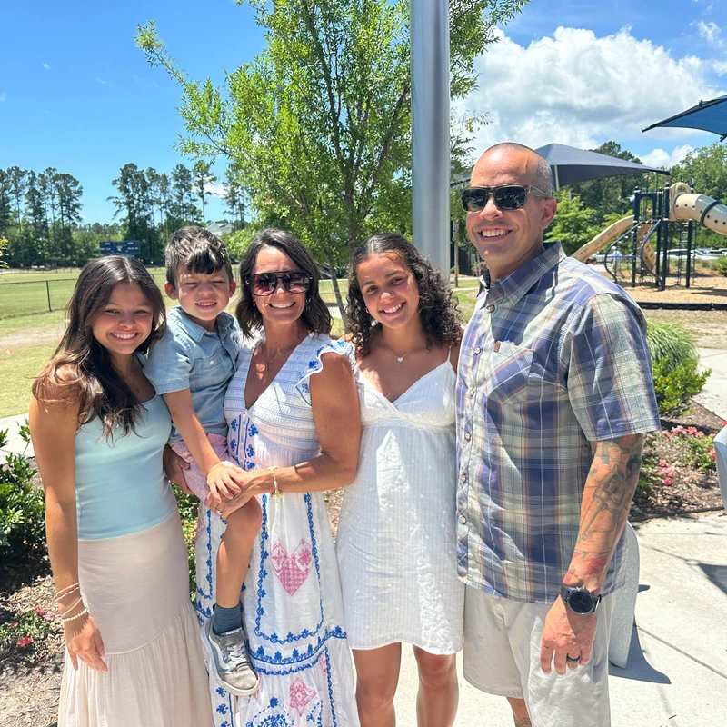 A family of five smiles for a photo outdoors on a sunny day, with a park and playground in the background.