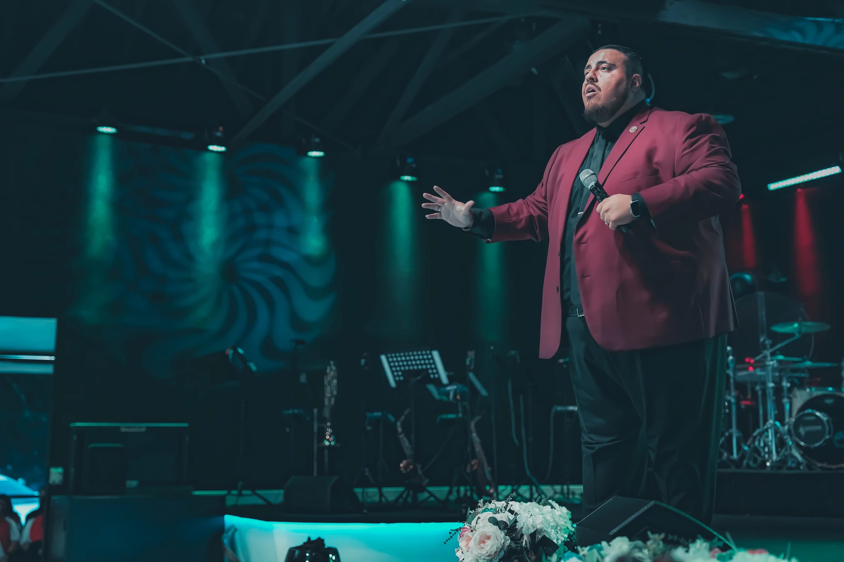 A man in a maroon jacket and black shirt stands on a dimly lit stage with green lights, speaking into a microphone with one hand outstretched.