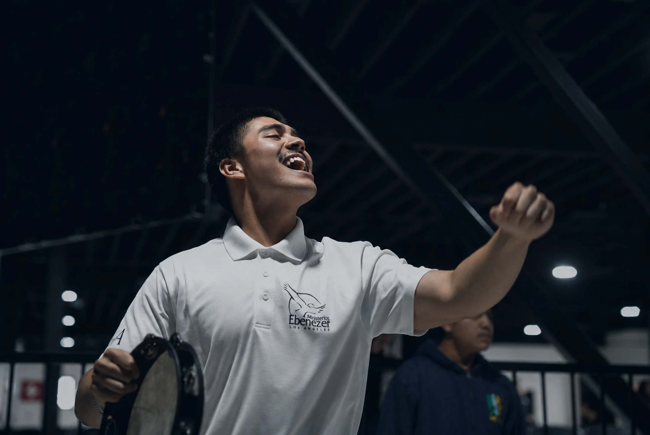 A young man in a white polo shirt sings with passion, looking upwards with his mouth open. He holds a tambourine in one hand and raises a fist in the air.