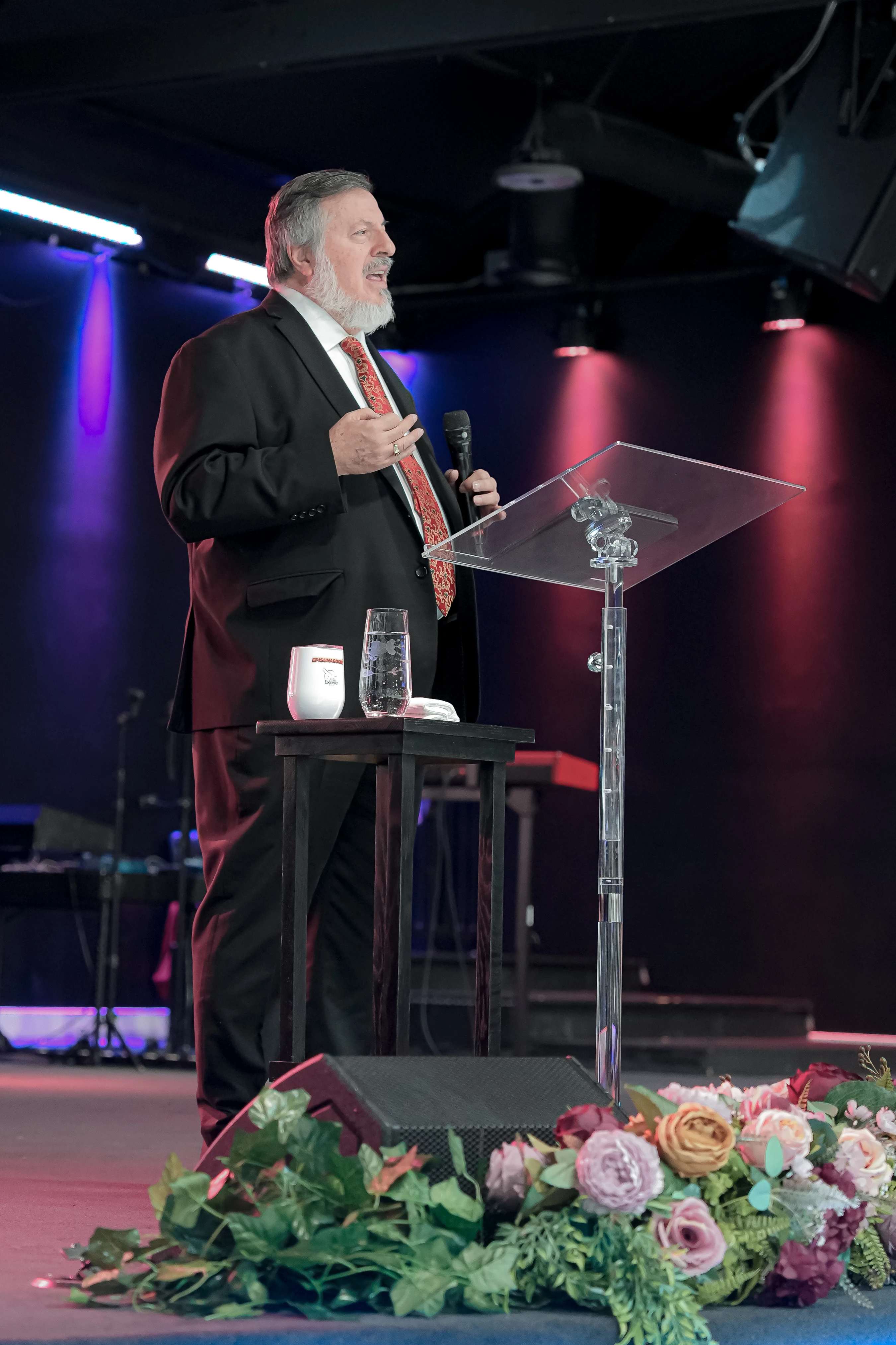 An older man with a gray beard, wearing a suit, speaks into a microphone while standing at a clear podium on a stage lit with purple and red lights.