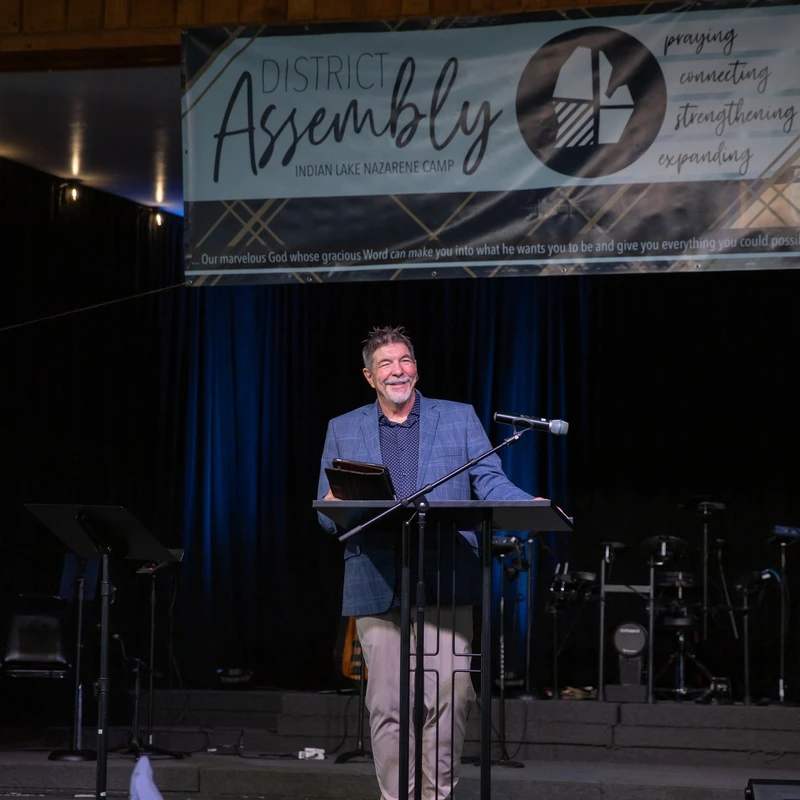 A smiling man in a blue blazer stands at a podium on a stage, speaking at the District Assembly at Indian Lake Nazarene Camp.