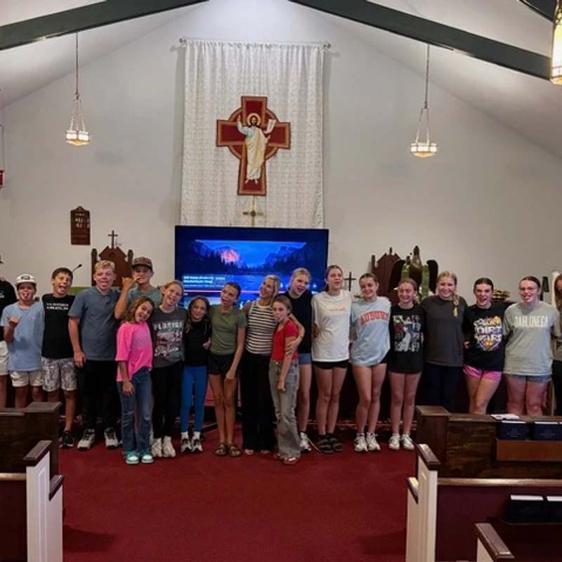 A large group of casually dressed young people, boys and girls, smiling for a group photo at the front of a church sanctuary.