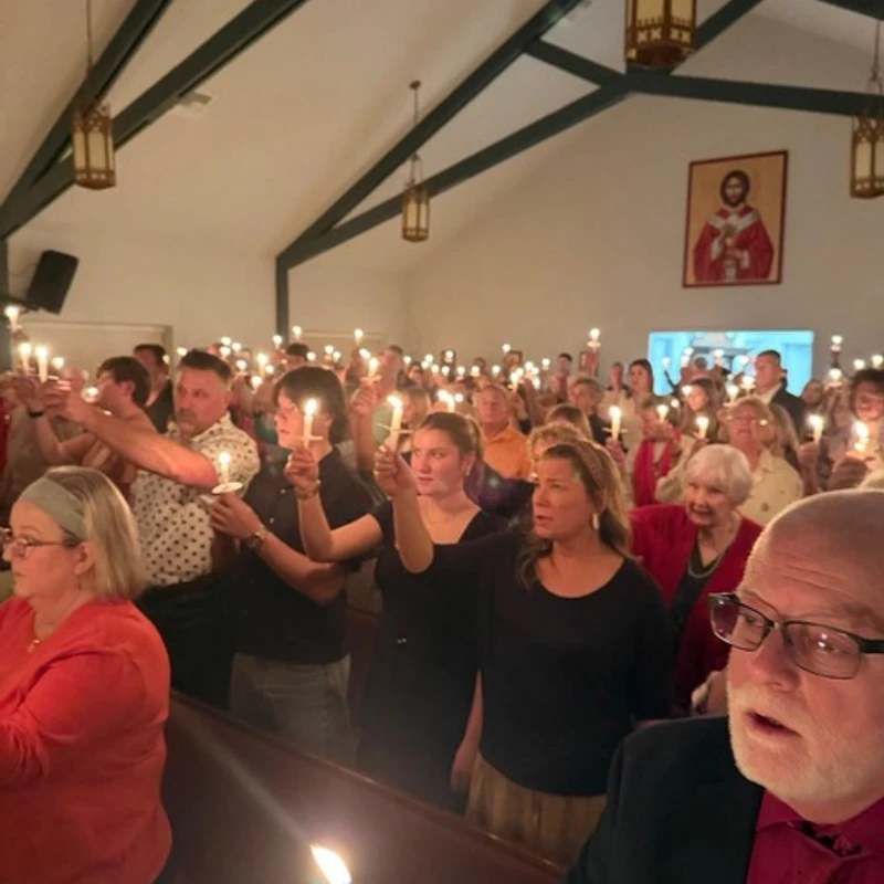 A diverse congregation stands in church pews, each person holding up a lit candle, filling the warmly lit room during a religious ceremony.