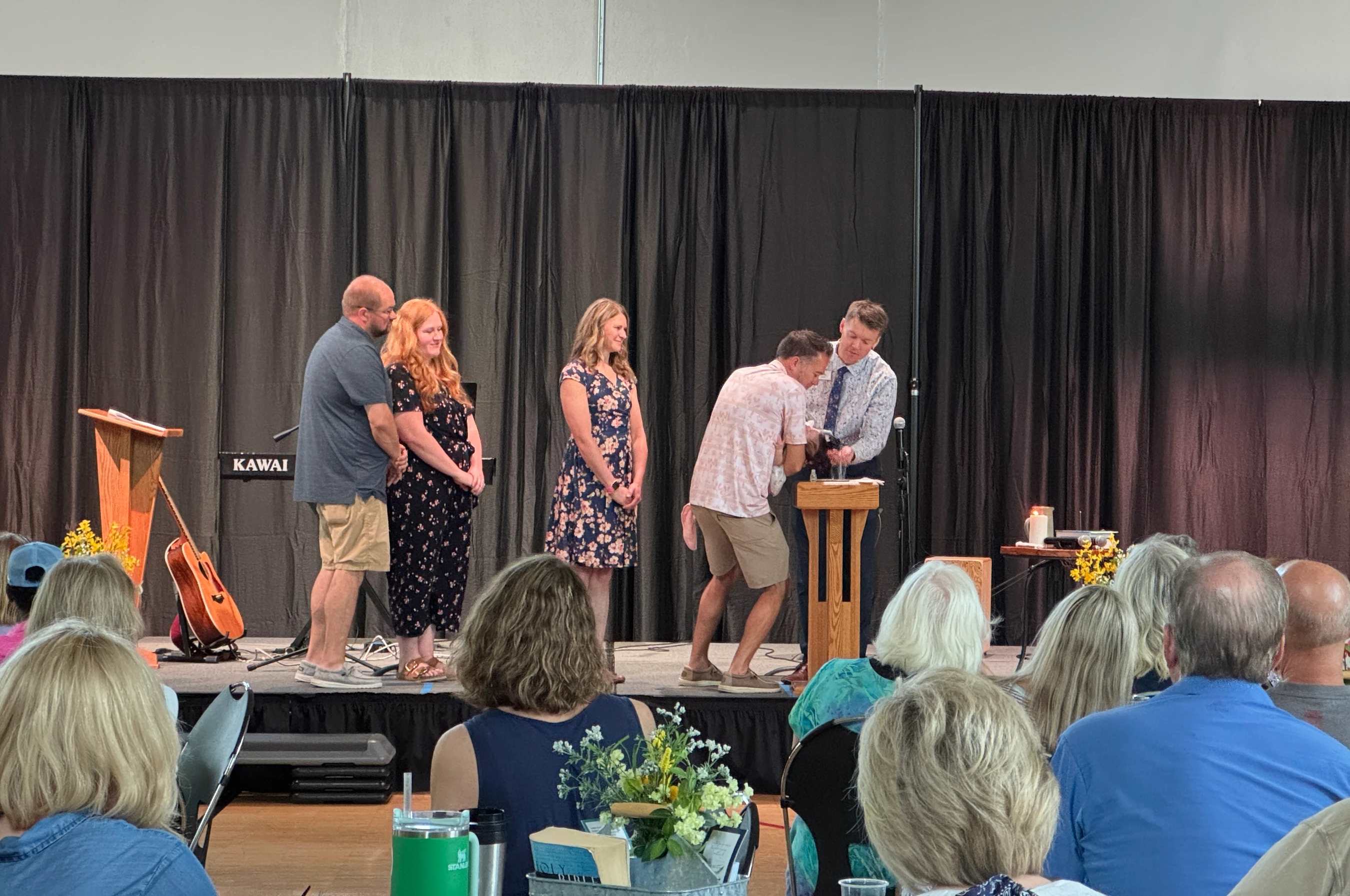 An audience watches as a pastor performs a ceremony, possibly a baptism or child dedication, for a family on a stage in front of a black curtain.