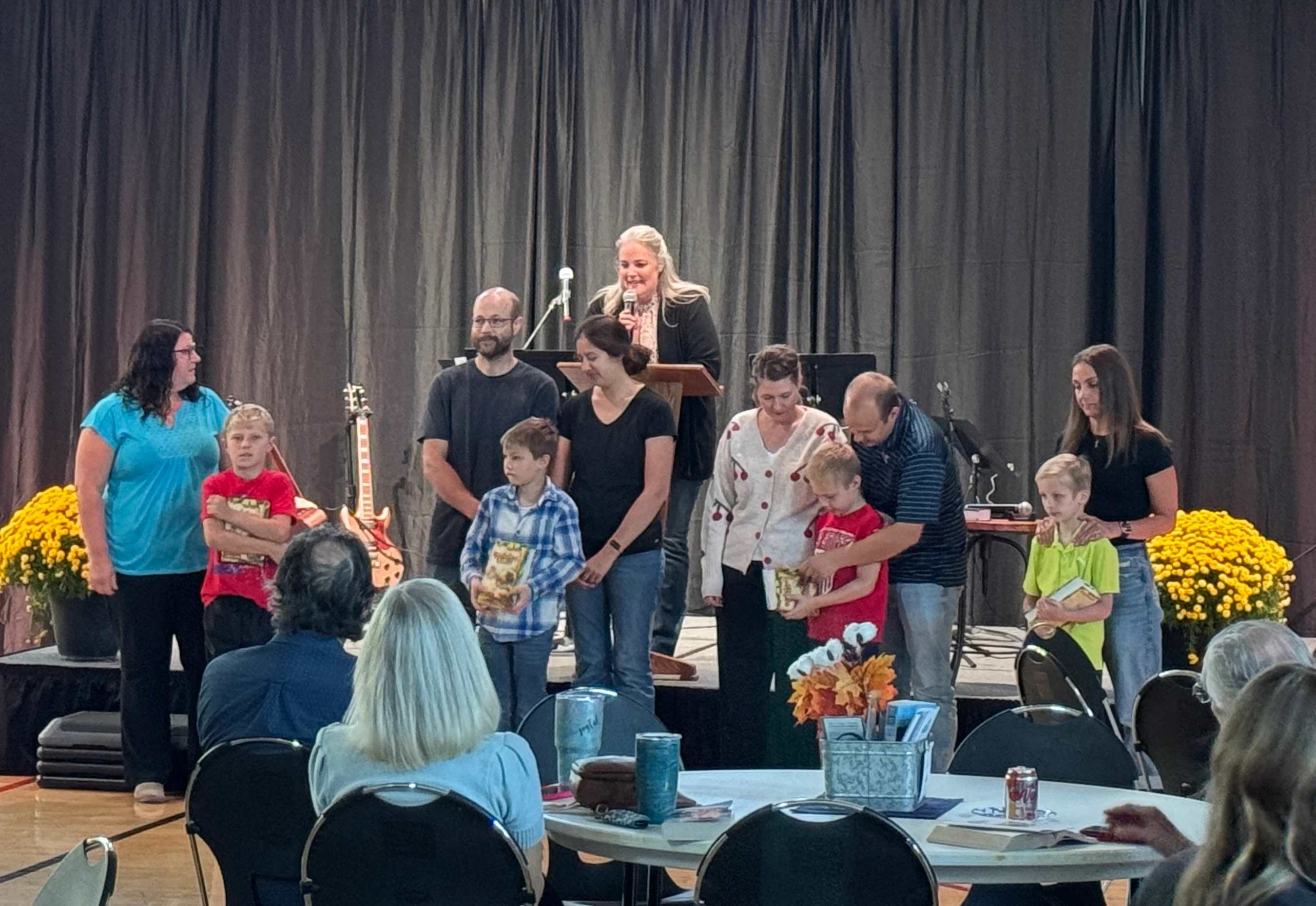 Several families with young children stand on a stage in front of a dark curtain. A woman at a podium speaks to an audience seated at tables.