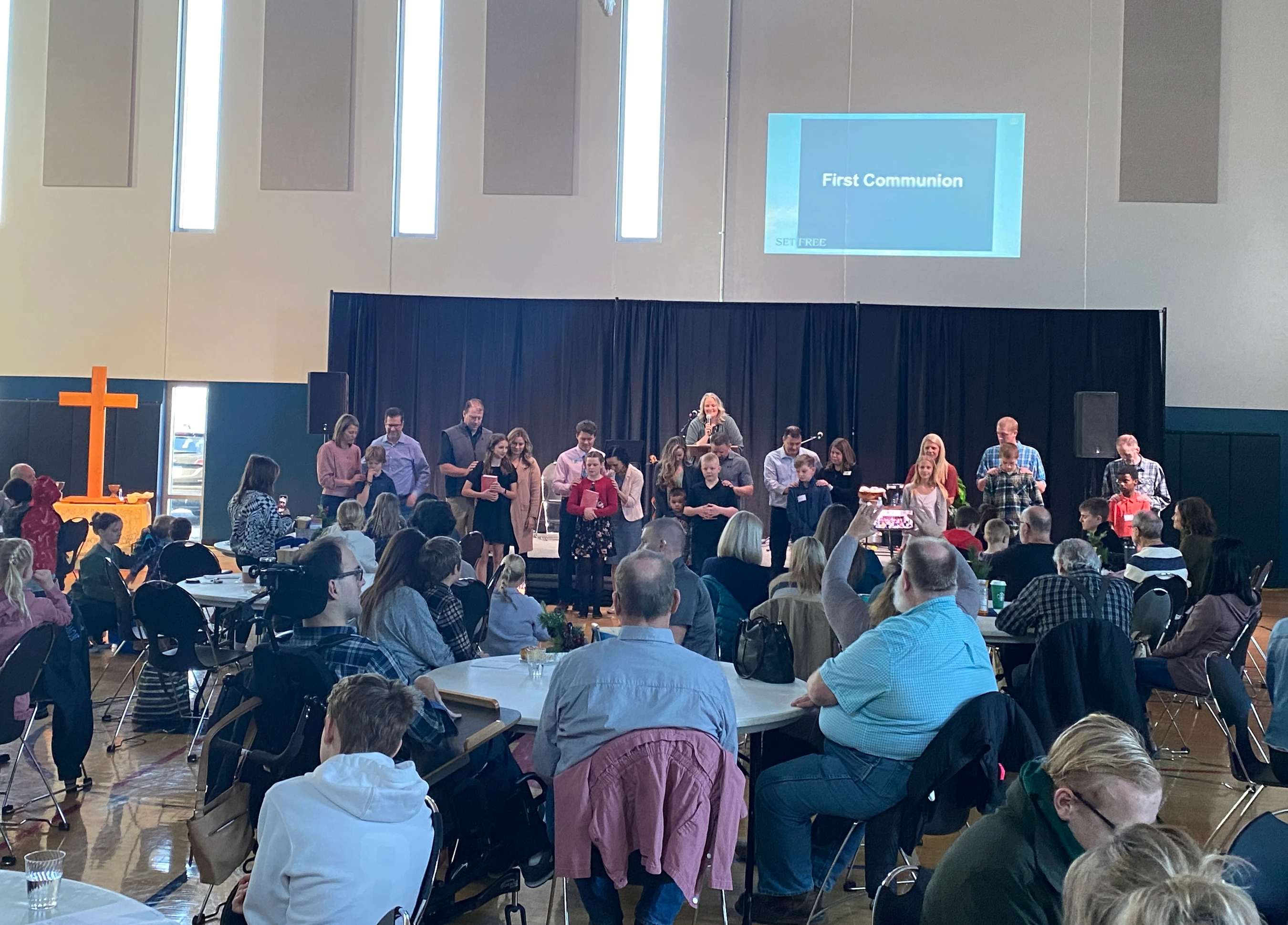 A large group of people are gathered in a hall, seated at round tables, watching a First Communion ceremony taking place on a stage at the front.