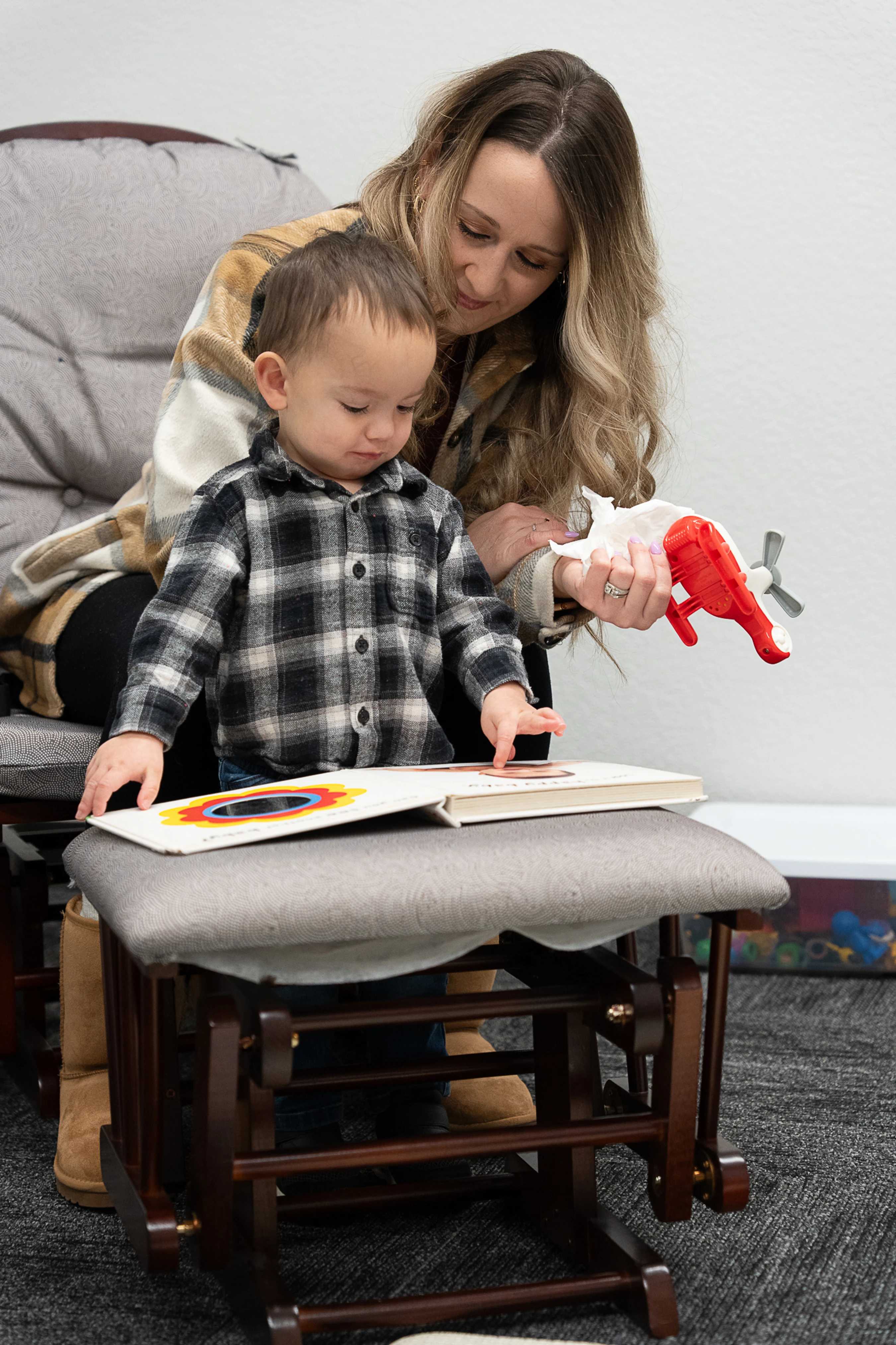 A woman sits in a rocking chair with her young son, who is standing on the footrest looking at a colorful picture book while she holds a toy plane.