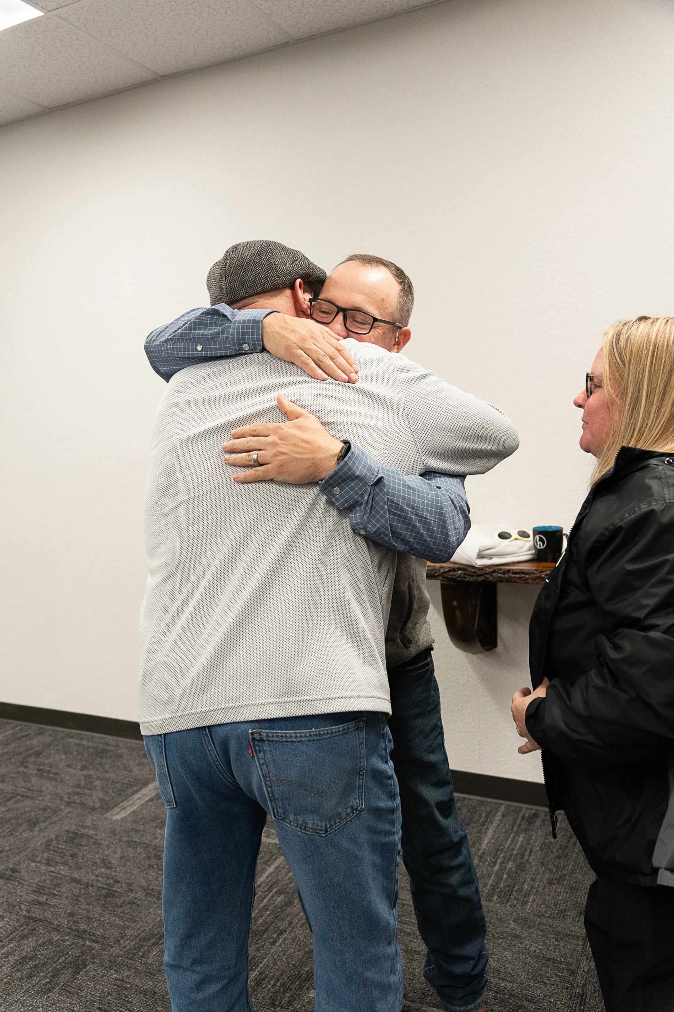 Two men hugging each other in a room with white walls and grey carpet. One wears a cap and plaid shirt, the other wears glasses. A woman looks on.