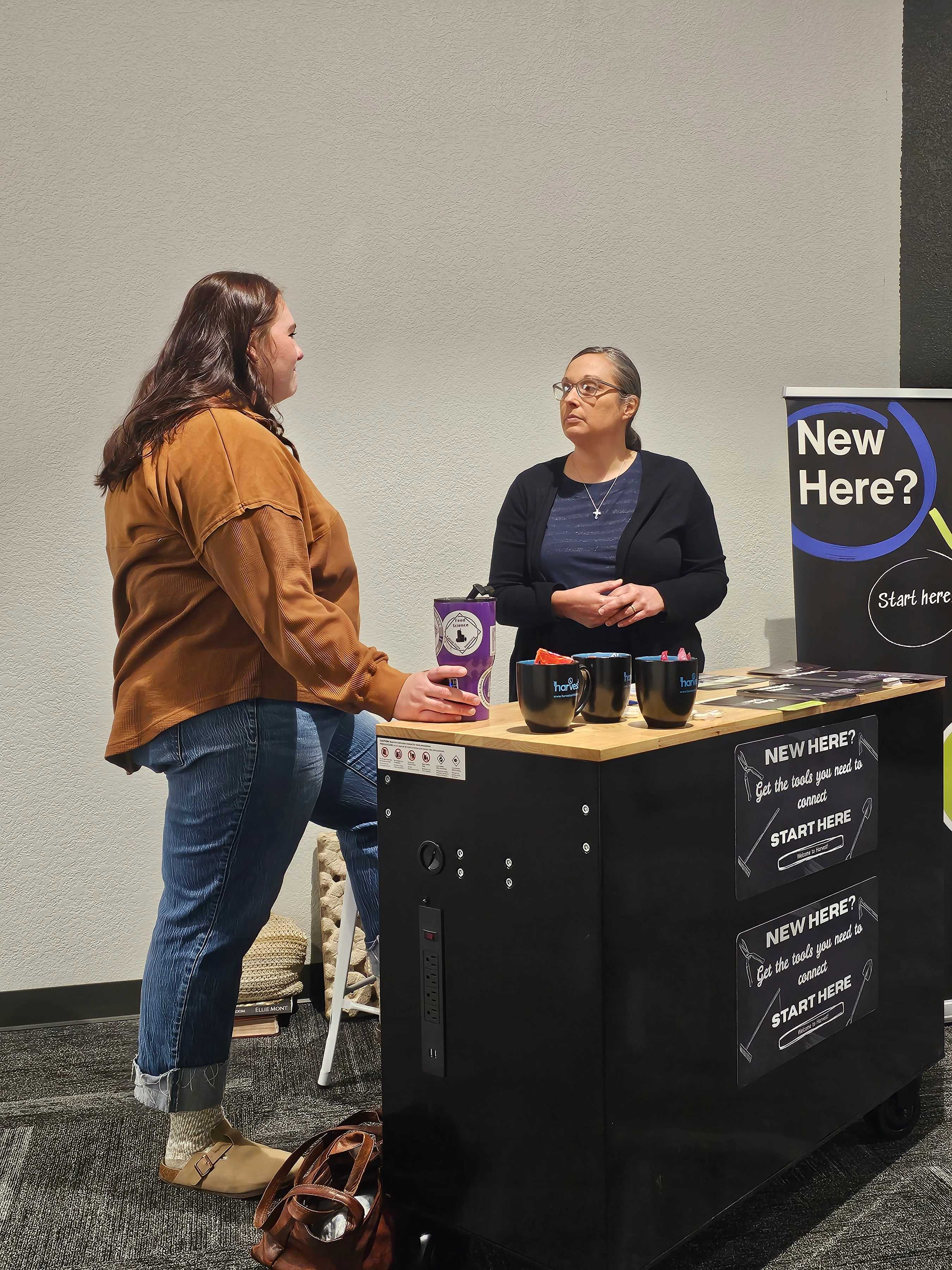 A young woman talks with a volunteer at a welcome booth. The booth has a sign that reads 'New Here? Start Here' to assist newcomers at an event.
