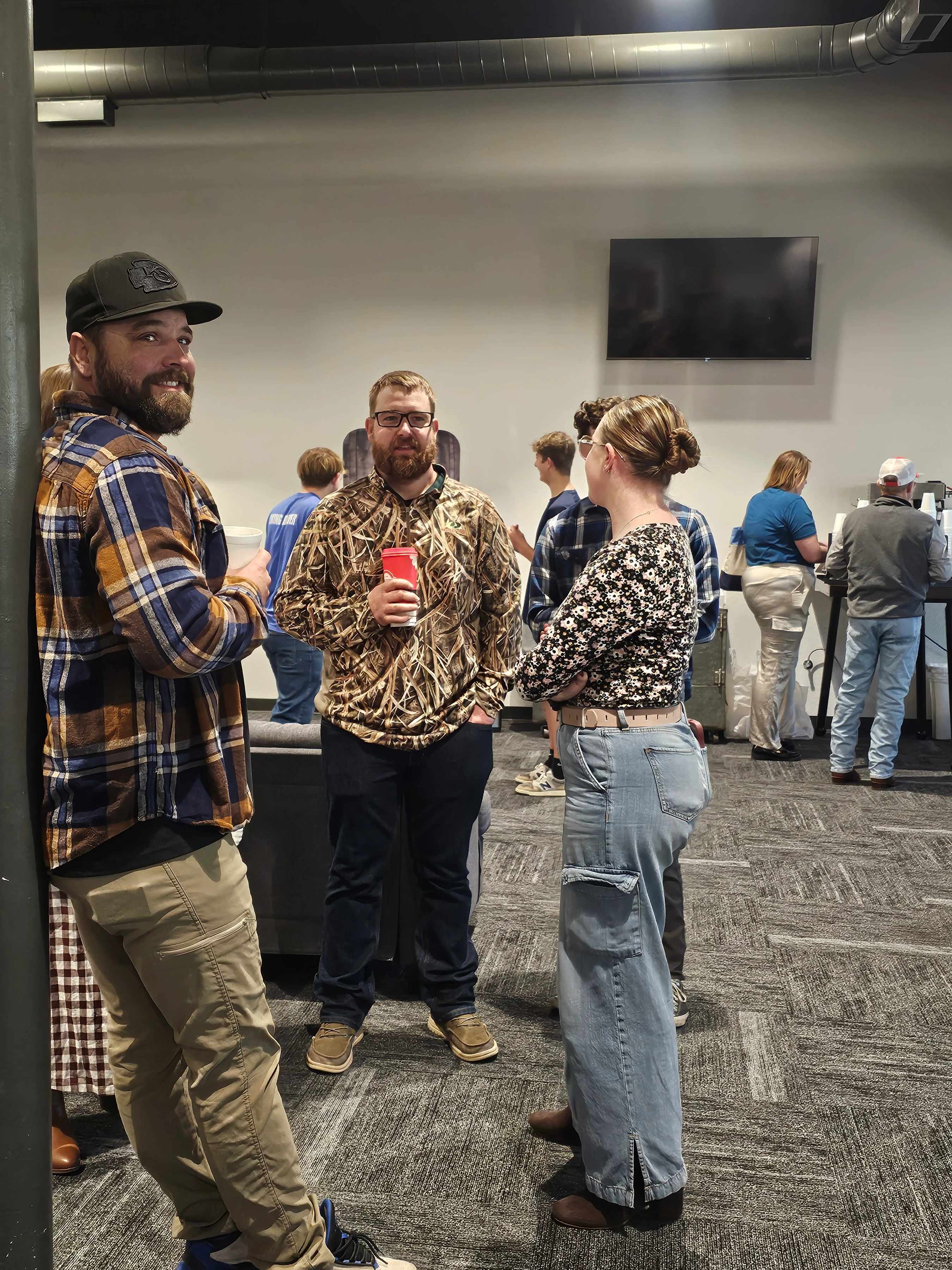 A man in a plaid shirt and cap smiles at the camera while standing with a man and woman who are talking at a casual indoor event with others in the background.