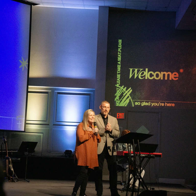 A woman speaks into a microphone while a man stands beside her on a dimly lit stage. A large screen behind them reads, 'Welcome, so glad you're here.'