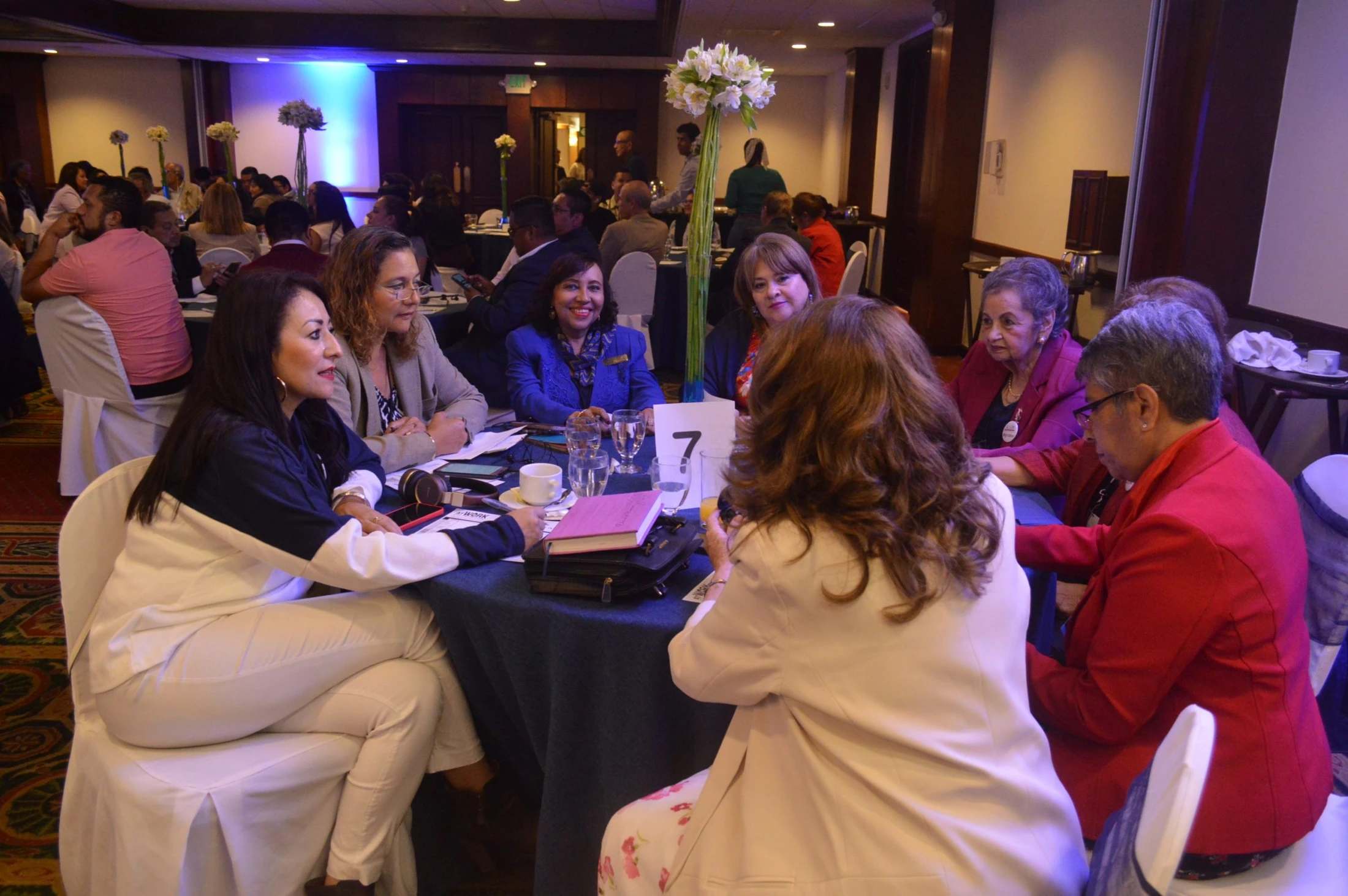 A group of women in professional attire are seated around a circular table with a blue tablecloth, actively talking during a conference or meeting.