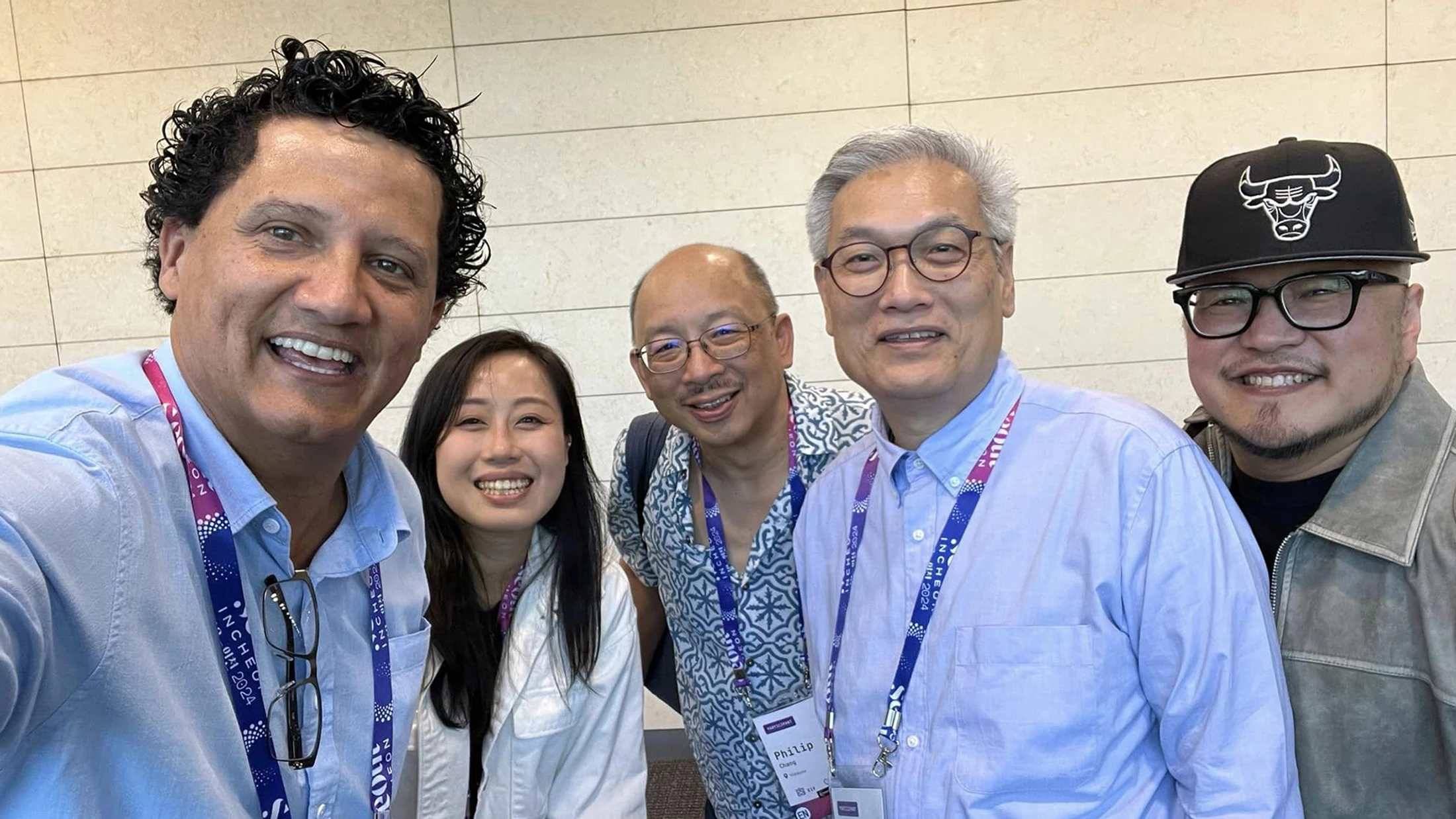 A group of five diverse people, one woman and four men, smile for a selfie indoors. They are all wearing lanyards for an event.