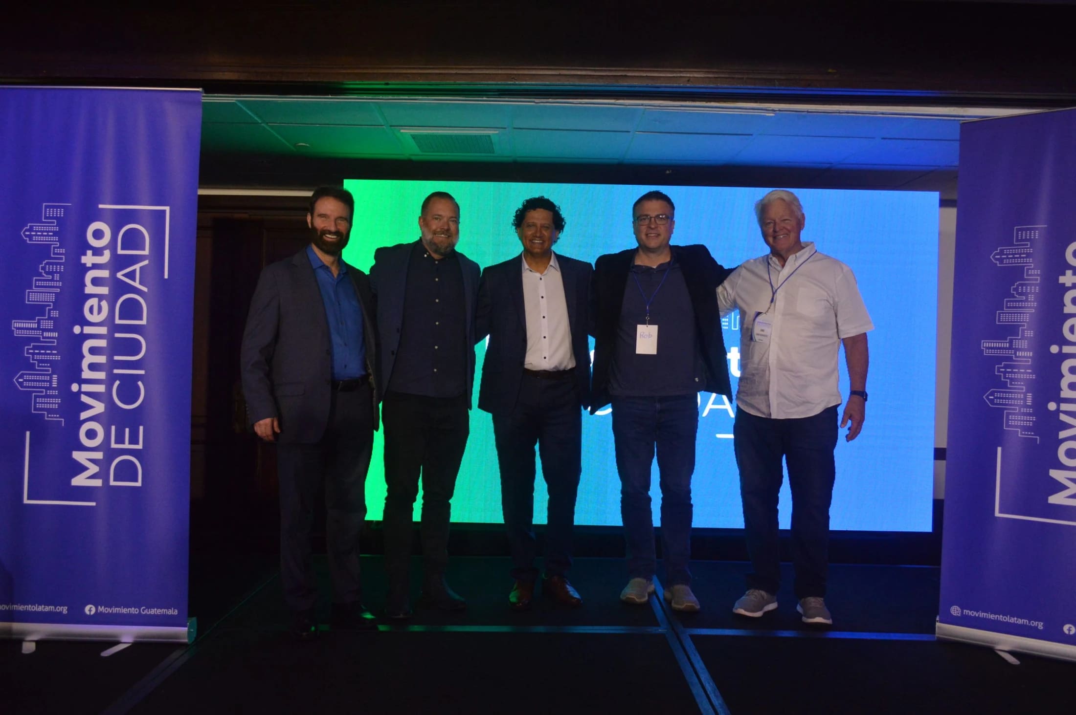 Five men in business casual attire stand together for a photo on a stage, flanked by blue banners for an event called 'Movimiento de Ciudad'.