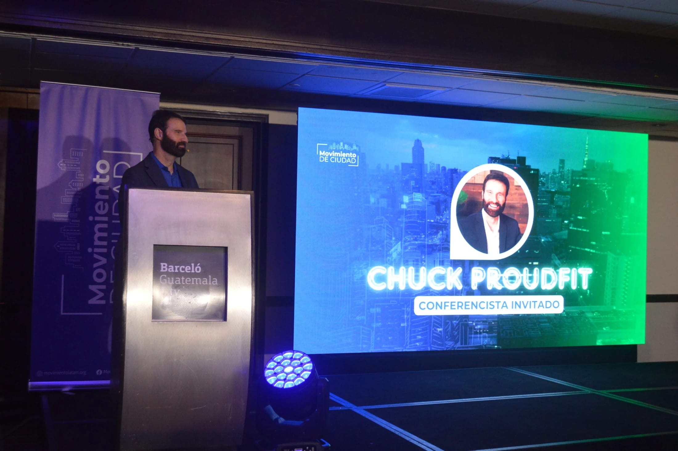 A man with a beard stands at a lectern on a stage. Behind him, a large screen shows his photo and name, Chuck Proudfit, identifying him as a guest speaker.
