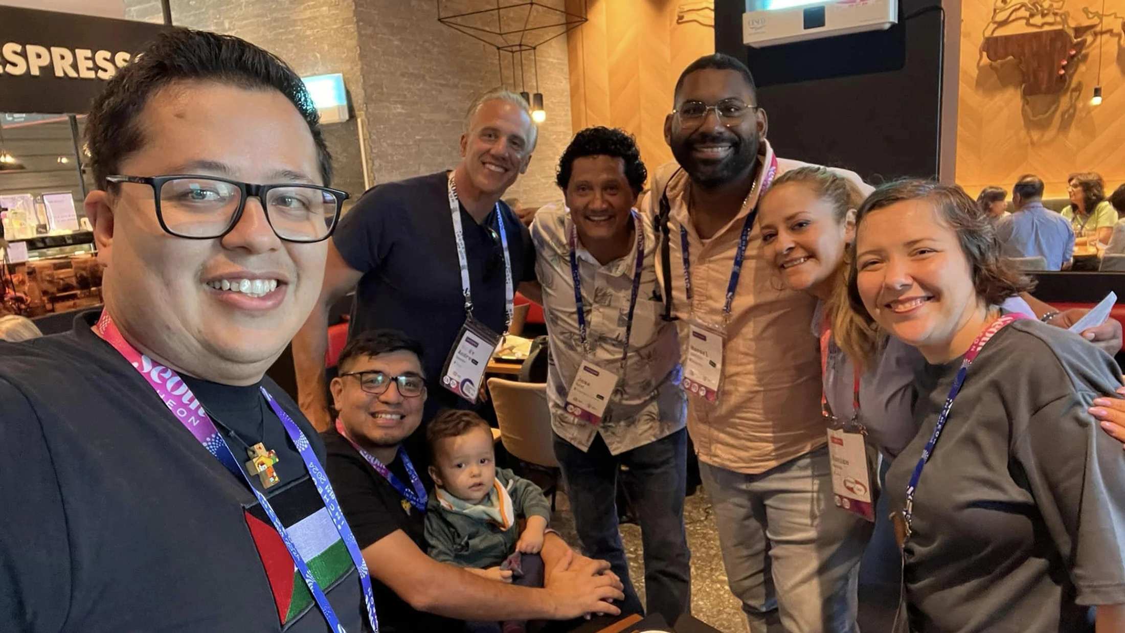 A diverse group of seven adults and a baby smile for a selfie taken in a cafe. They are all wearing event lanyards and look happy.
