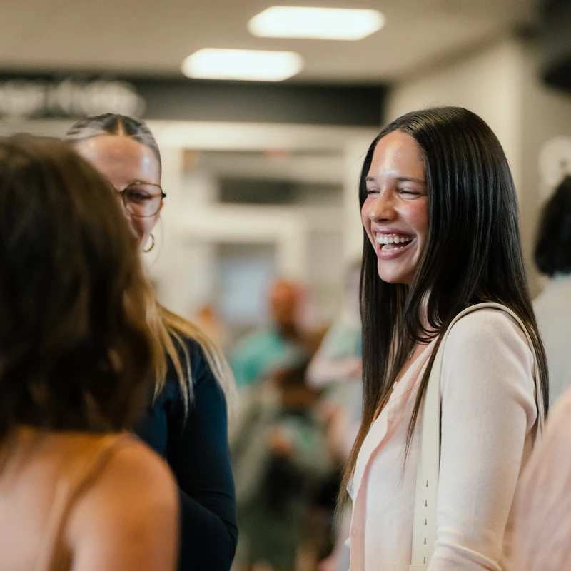 A young woman with long dark hair laughs with her eyes closed while talking to friends in a brightly lit indoor space.