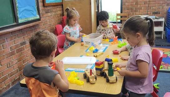 Four young children sit and stand around a long table in a classroom, playing with colorful building blocks and wooden train toys.