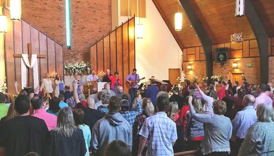 A view from the back of a church sanctuary filled with people standing and singing, some with hands raised, as a worship band performs on stage.
