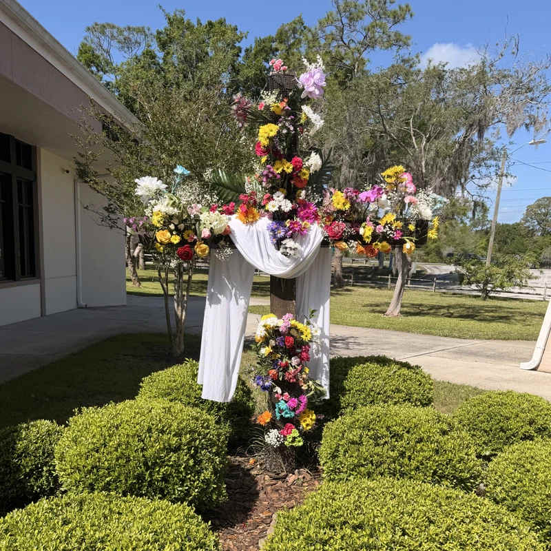 A wooden cross stands outdoors in a garden, covered in vibrant, multicolored flowers and draped with a white sash under a clear blue sky.