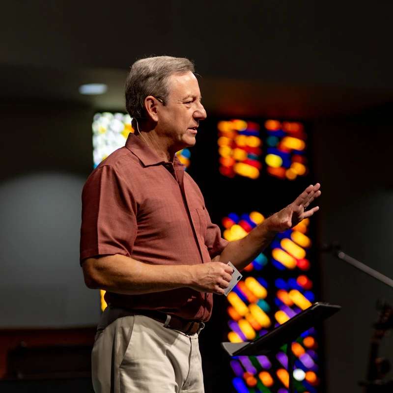 A middle-aged man in a reddish-brown shirt stands at a lectern, speaking and gesturing. A colorful, out-of-focus stained-glass window is behind him.