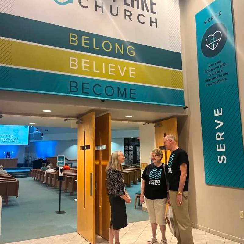 Three people stand and talk in a church lobby under a large sign for East Lake Church that reads 'Belong, Believe, Become'.