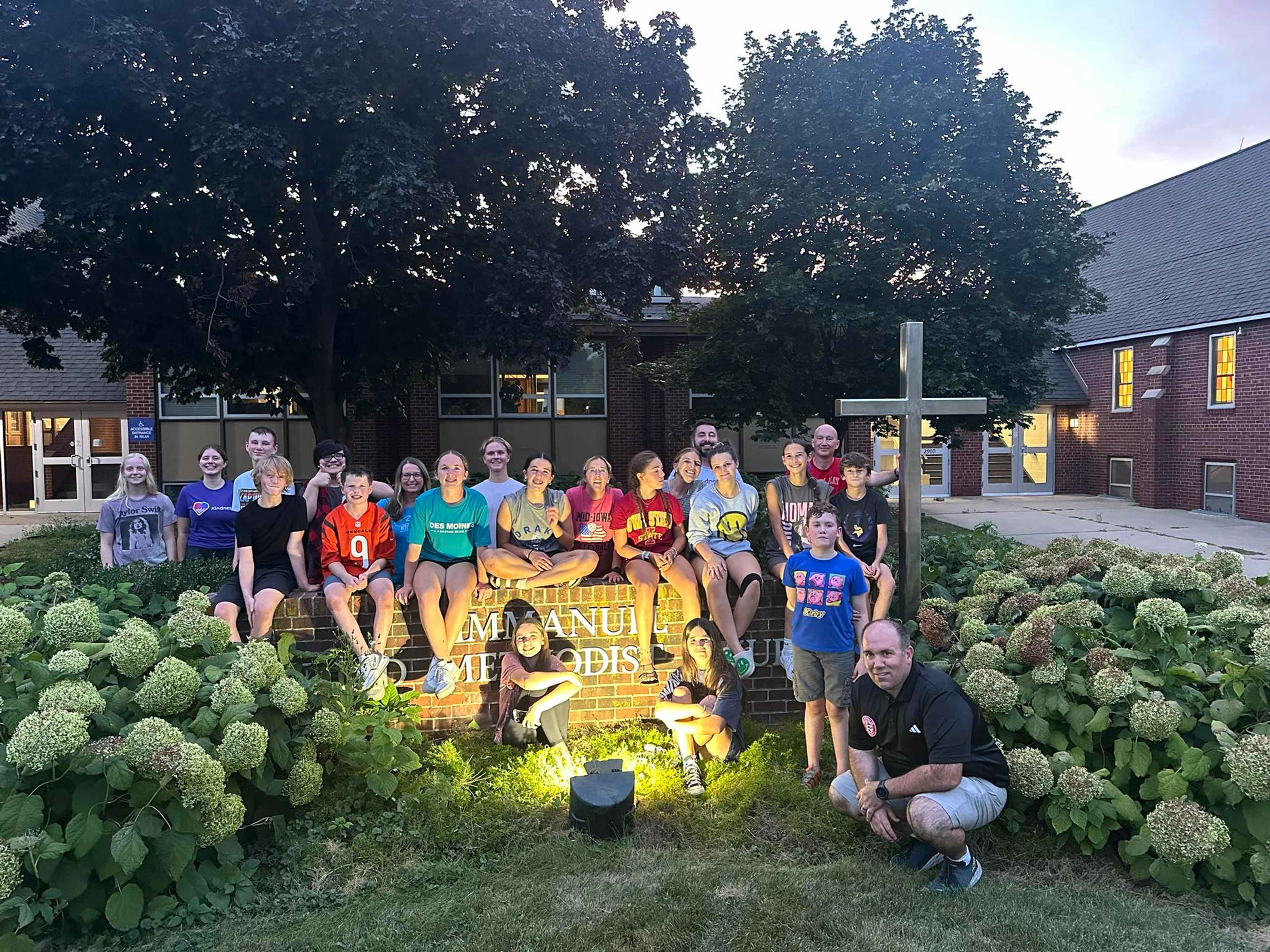 A large group of teenagers and a few adults smile for a photo outdoors in the evening in front of a brick church building and a large cross.