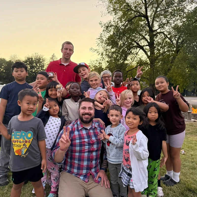 A large, diverse group of smiling children and two men pose for a photo on a lawn at dusk. Many are making peace signs with their hands.
