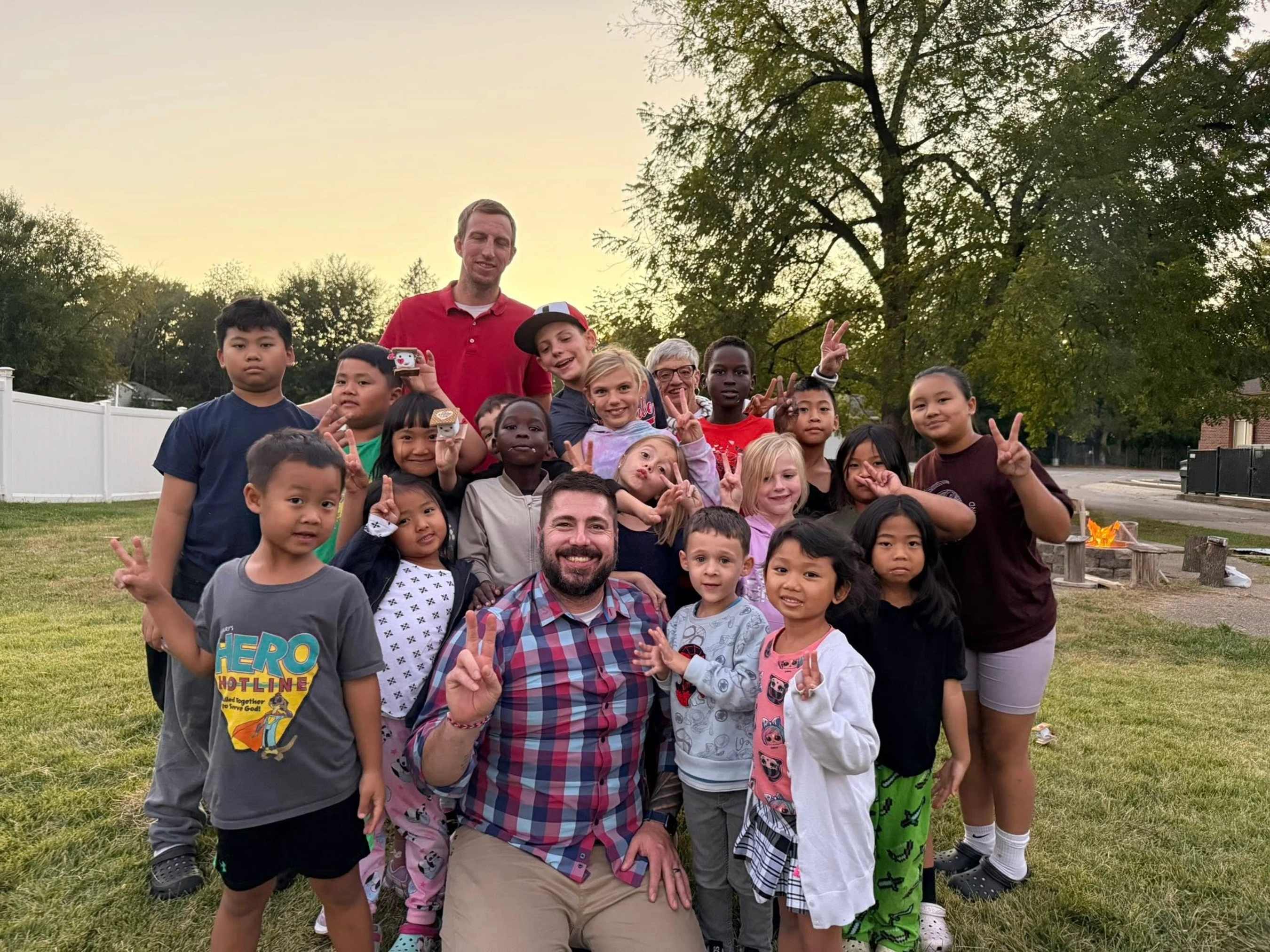 A large, diverse group of smiling children and two men pose for a photo on a lawn at dusk. Many are making peace signs with their hands.