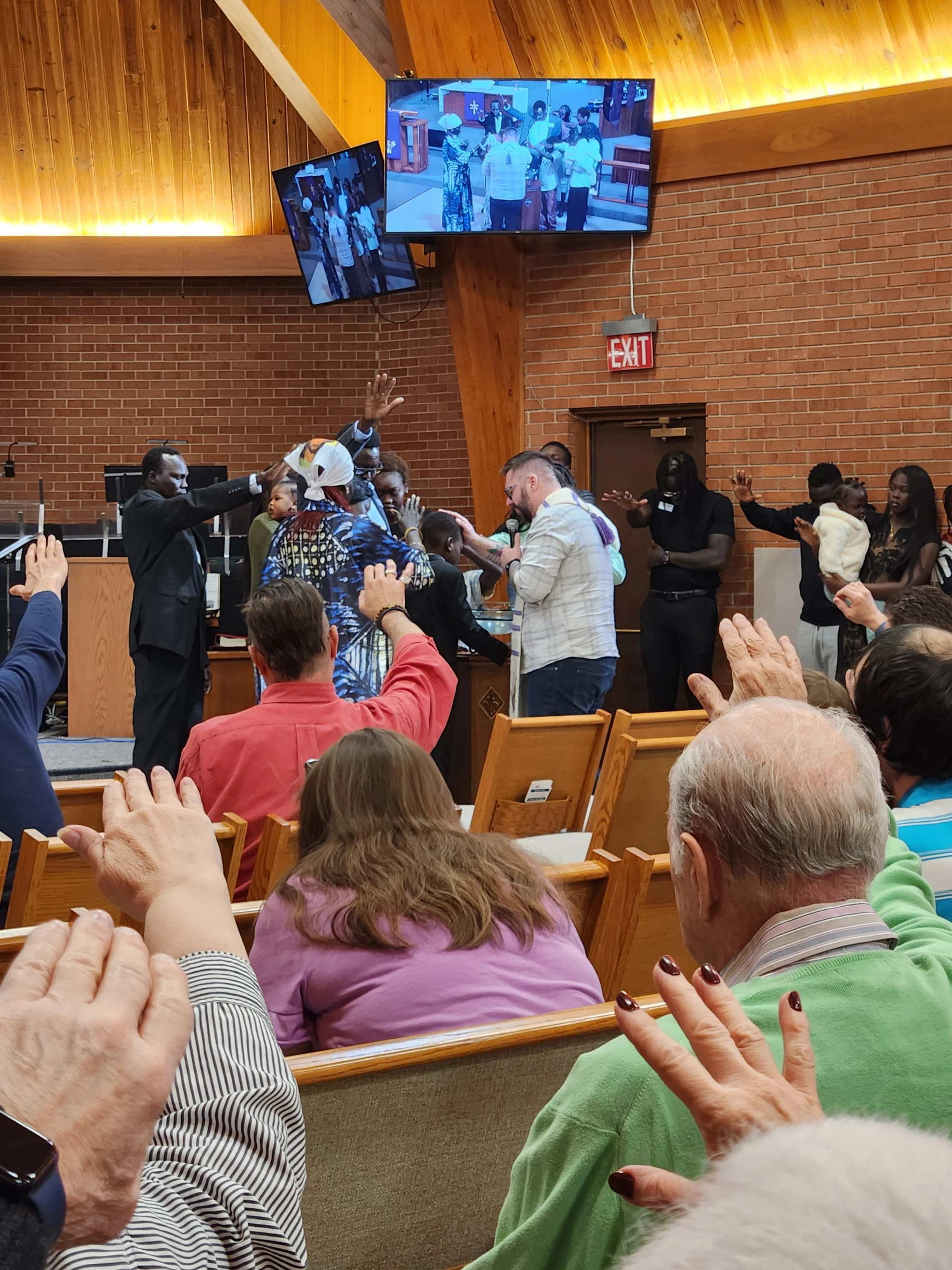 A diverse congregation in a church with hands raised in prayer towards people on stage receiving a blessing during a service.